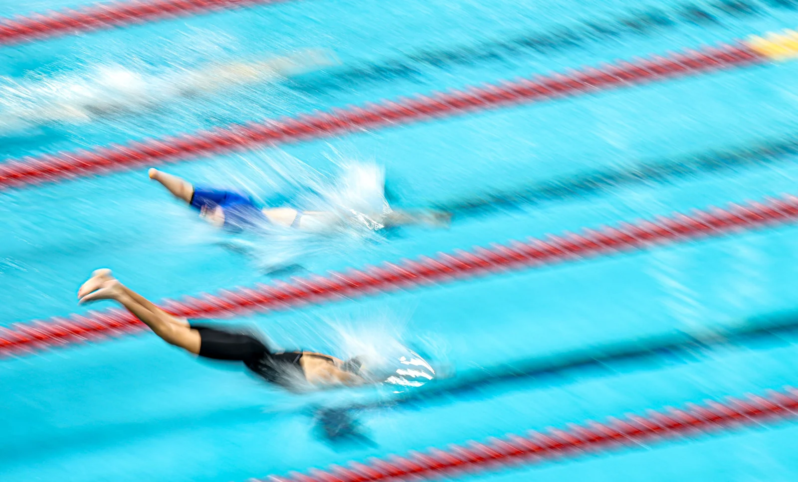 Nadadores competem em raias de uma piscina; a fotografia acompanha os movimentos dos braços sobre a água.