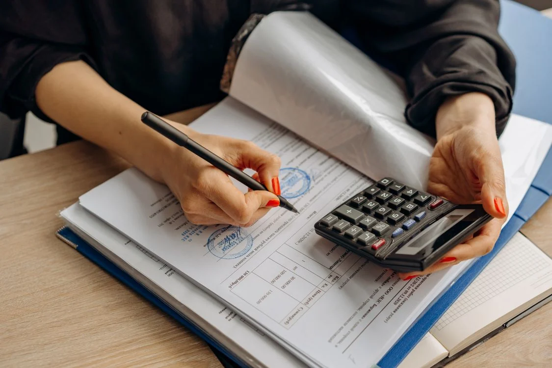 Mãos de uma pessoa analisam documentos sobre uma mesa enquanto utilizam uma calculadora.