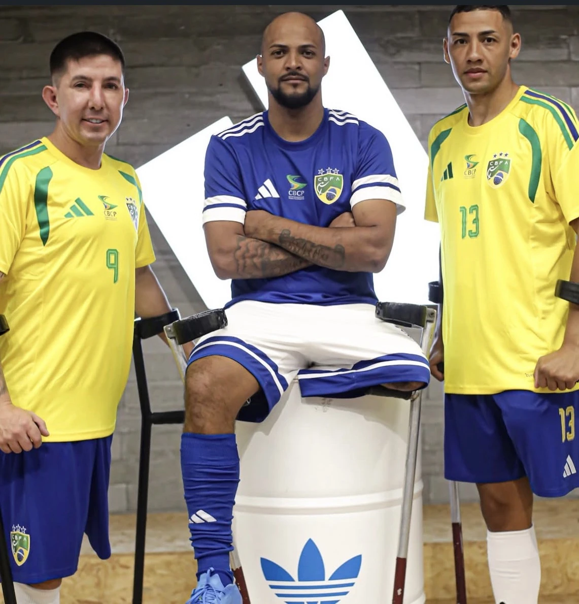 Três pessoas em uniformes de futebol da Adidas, duas de amarelo e uma de azul, posam com muletas; a pessoa ao centro está sentada sobre um tambor branco.