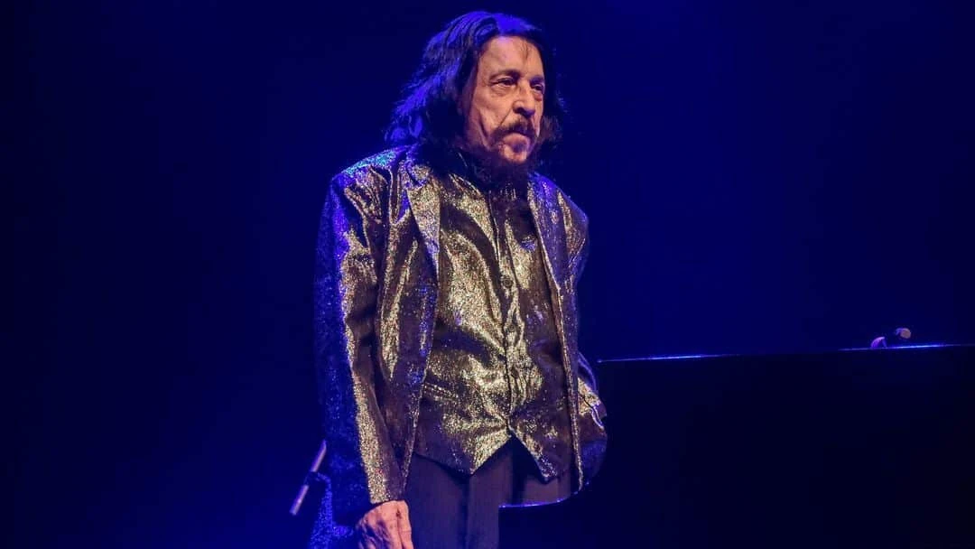 Benito di Paula em um palco com iluminação azul, vestindo paletó e colete dourados, ao lado de um piano preto.