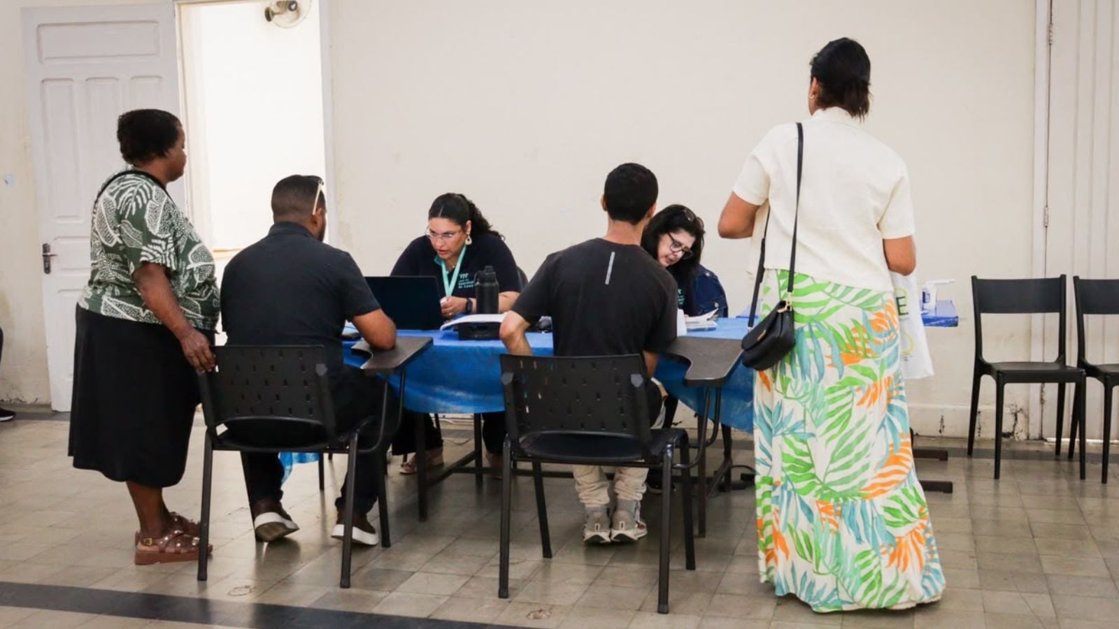 Duas profissionais atendem pessoas sentadas diante de uma mesa azul, com computador e documentos; outras duas pessoas aguardam em pé.