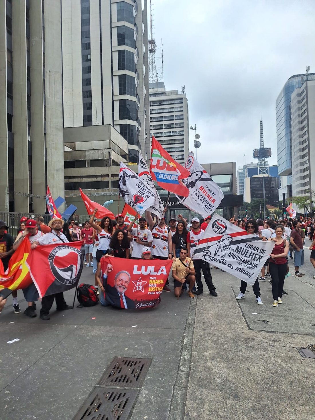 Grupo de manifestantes reunido na Avenida Paulista segura faixas e numerosas bandeiras, entre elas símbolos de torcidas e uma faixa vermelha de apoio a Lula. Prédios altos ladeiam a via sob céu nublado.