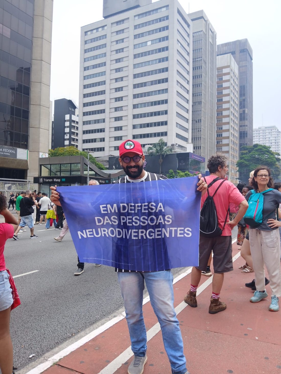 Thiago Almeida sorri, de boné vermelho, óculos, camisa do Corinthians e calça jeans, segurando uma faixa azul com a frase “Em defesa das pessoas neurodivergentes”. Ao fundo, aparecem manifestantes, prédios e o acesso à estação Trianon-Masp.
