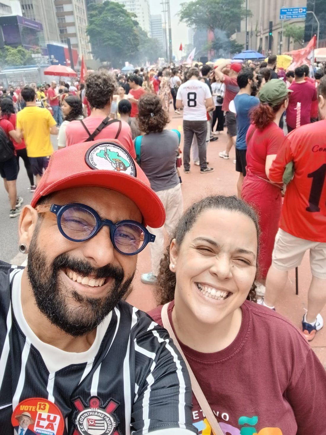 Selfie de Thiago Almeida, de barba, óculos azuis, boné vermelho e camisa do Corinthians, ao lado de sua esposa, Regiane Soares, de camiseta vinho. Ambos sorriem; atrás deles, manifestantes ocupam a avenida.