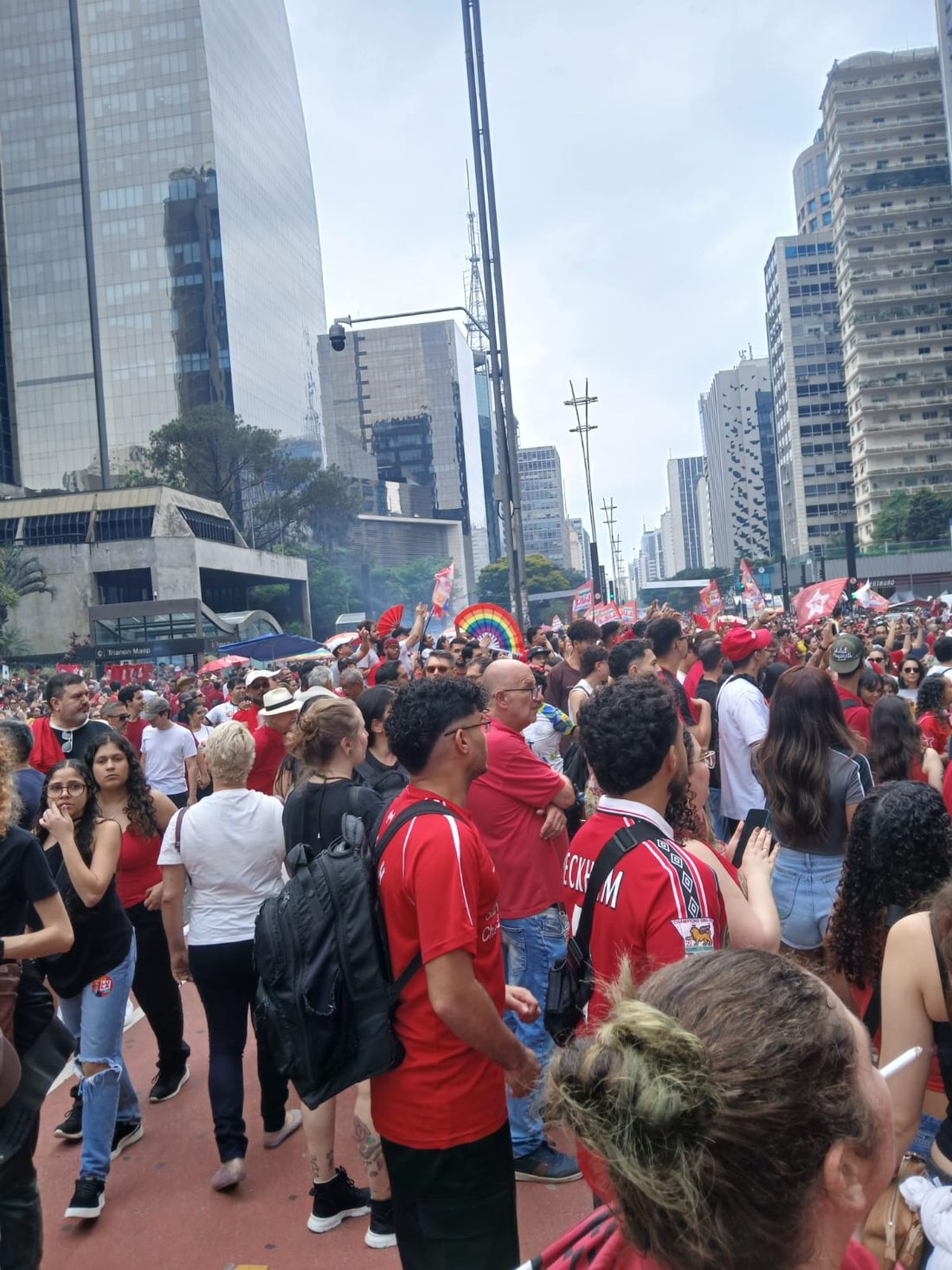 Vista ao nível da rua de uma multidão entre prédios da Avenida Paulista. Pessoas usam roupas de diferentes cores, incluindo camisas de futebol; bandeiras aparecem ao fundo.