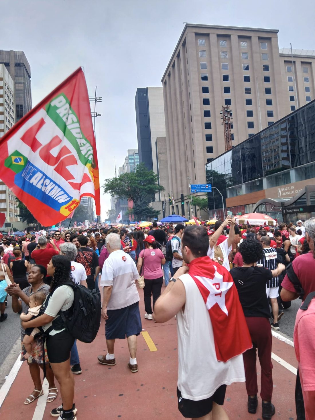 Manifestantes caminham entre prédios altos. À esquerda, uma grande bandeira vermelha traz o nome Lula. Em primeiro plano, uma pessoa veste uma bandeira vermelha e branca sobre os ombros.
