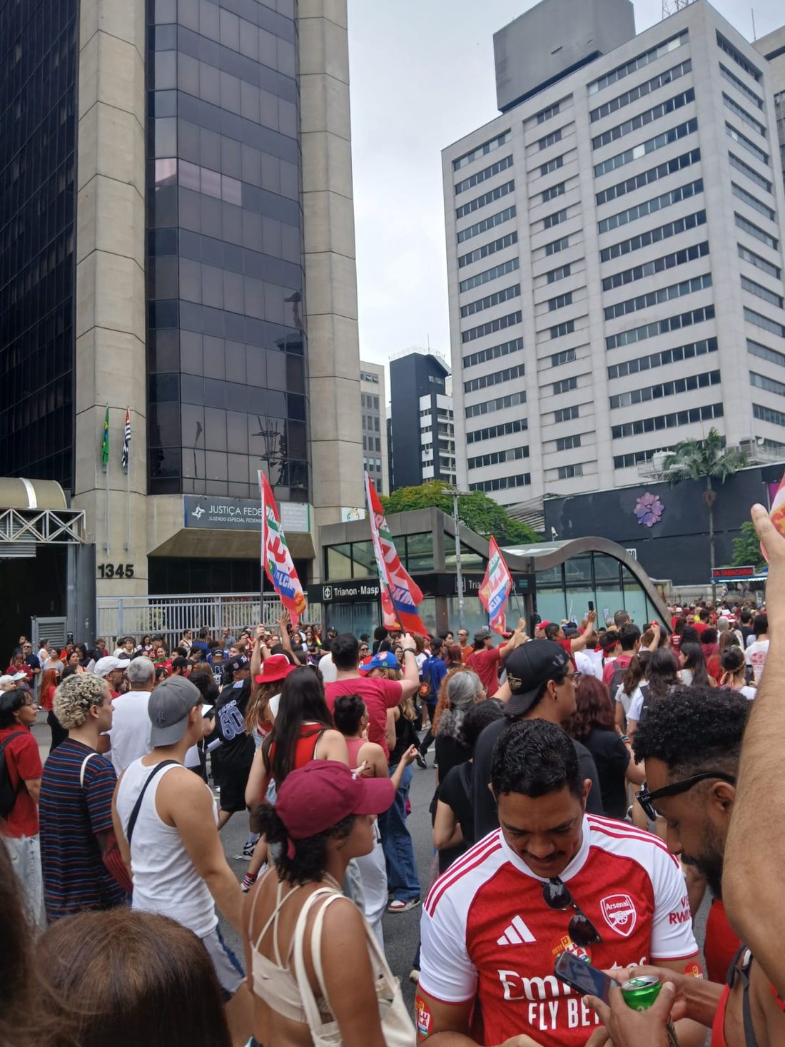 Manifestantes se concentram em uma avenida cercada de prédios altos. Bandeiras vermelhas aparecem acima do grupo; em primeiro plano, à direita, um homem usa camisa vermelha de futebol.