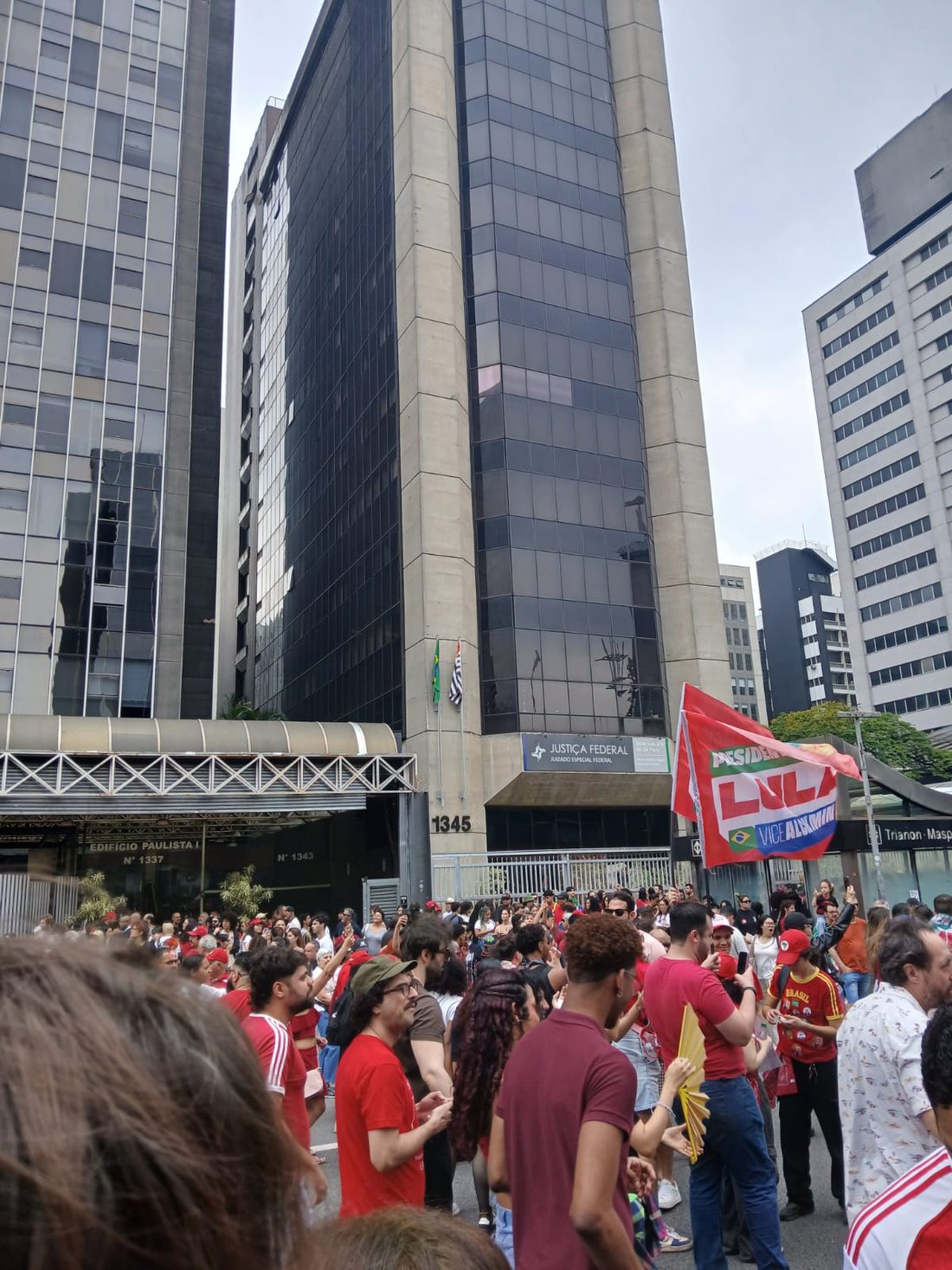 Grupo de manifestantes diante de edifícios altos com fachadas de vidro e concreto. Uma bandeira vermelha com o nome Lula se destaca à direita.
