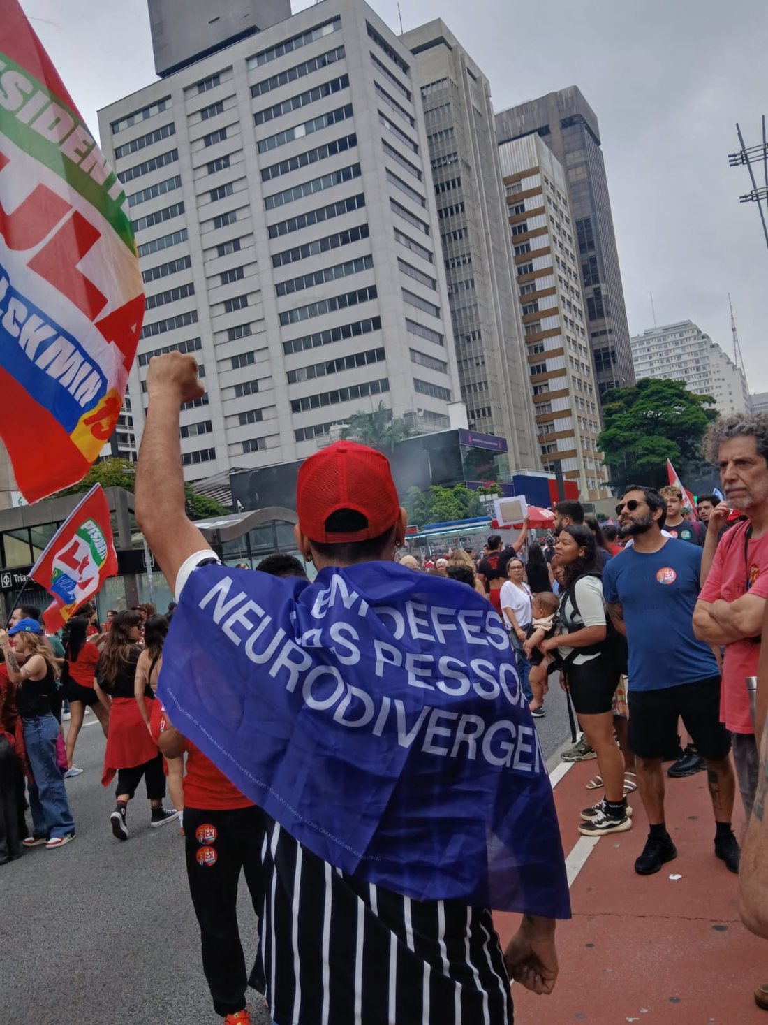 Participante de costas, com boné vermelho e camisa listrada em preto e branco, usa uma faixa azul sobre os ombros. Parte da mensagem em defesa das pessoas neurodivergentes está visível; há manifestantes e prédios ao fundo.