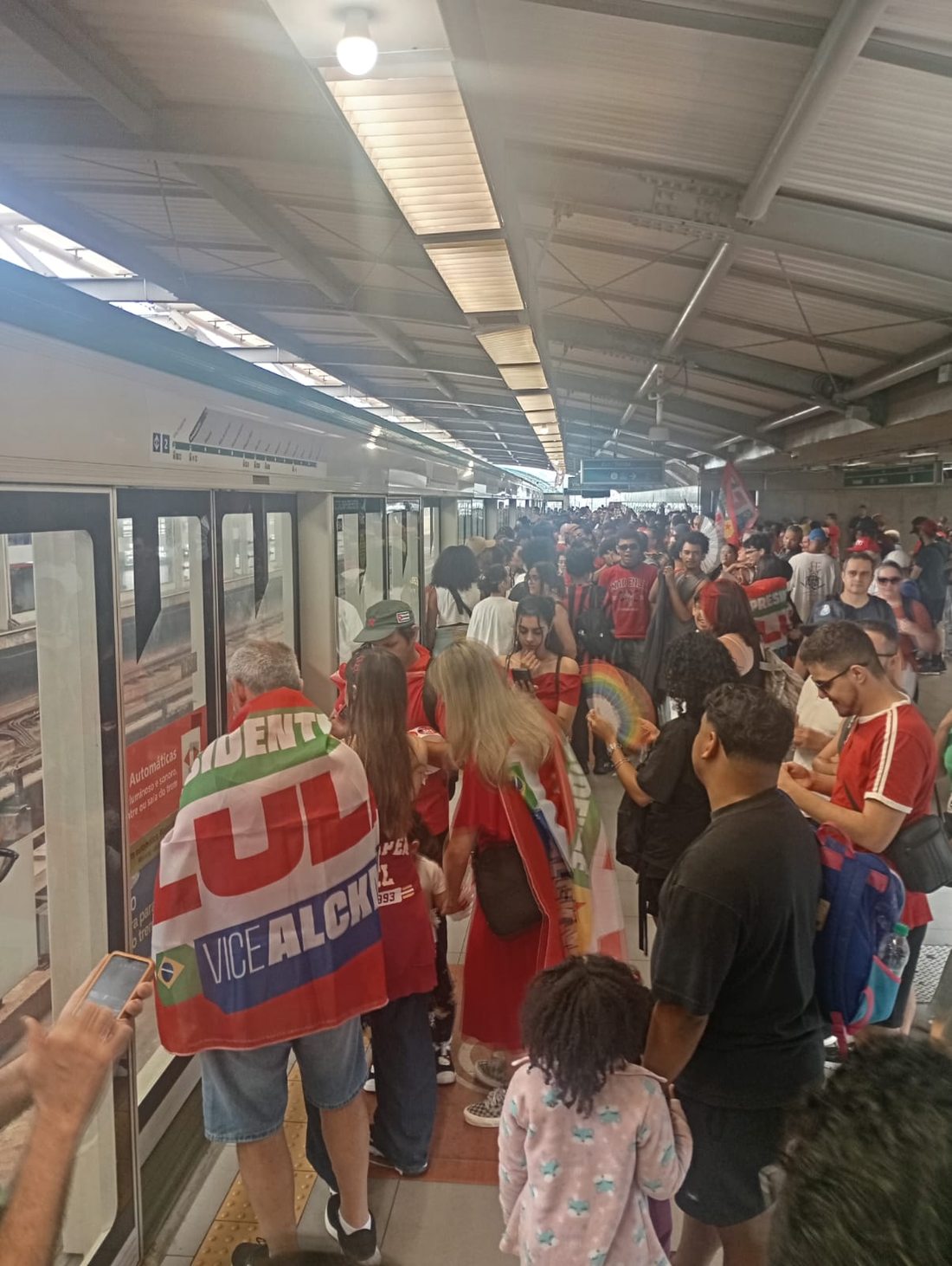 Plataforma coberta e cheia de pessoas ao lado de um trem com portas abertas. No centro, uma pessoa carrega uma bandeira de campanha vermelha com os nomes Lula e Alckmin.