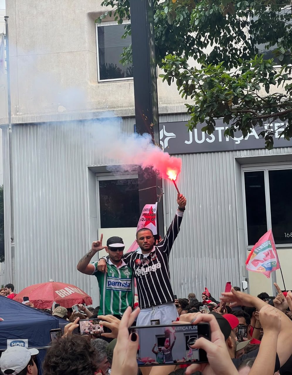 Dois homens em posição elevada seguram sinalizadores que produzem fumaça clara e vermelha. Um usa camisa verde e o outro, camisa listrada em preto e branco. Pessoas abaixo registram a cena com celulares.