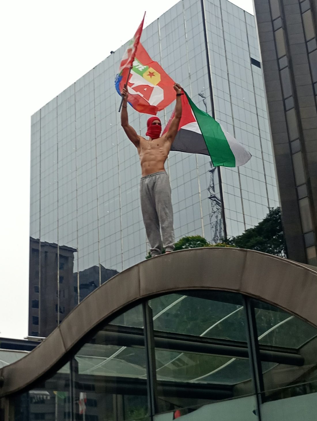 Pessoa sem camisa, em cima da cobertura curva de uma estrutura de vidro, ergue bandeiras vermelha e verde. Uma grande fachada cinza ocupa o fundo da fotografia.