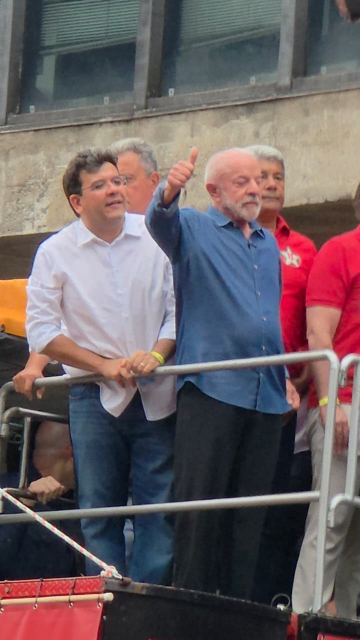 Lula, de camisa azul, ergue o braço em uma plataforma com grades, ao lado de outras pessoas, diante de uma fachada de concreto.