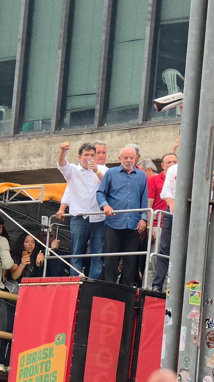 Vista vertical de uma plataforma com grades e painel vermelho. Lula, de camisa azul, aparece junto de outros participantes; ao lado, um homem de camisa clara ergue o braço.