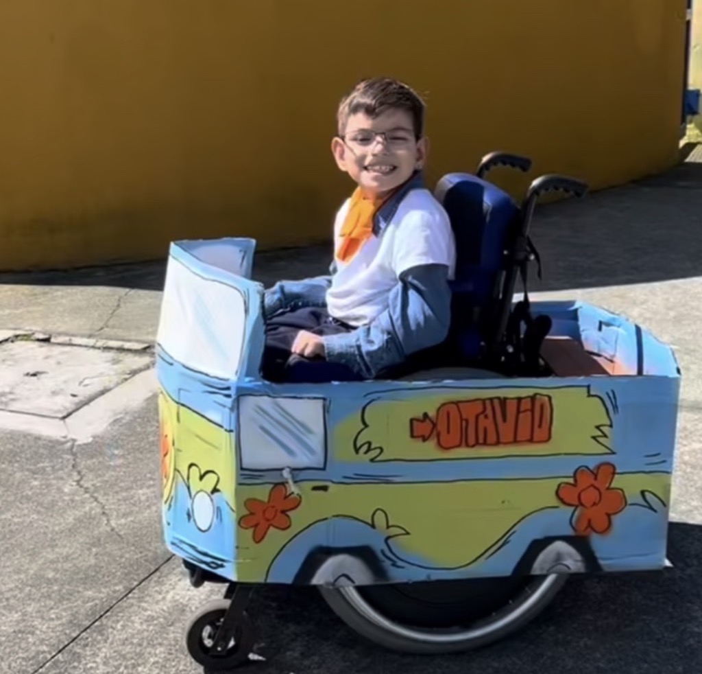 Otávio sorri de óculos, blusa branca sobre camisa azul e lenço laranja, em cadeira de rodas decorada como a van azul e verde de Scooby-Doo, com flores laranjas e o nome Otávio na lateral. Ao fundo, há um muro amarelo.