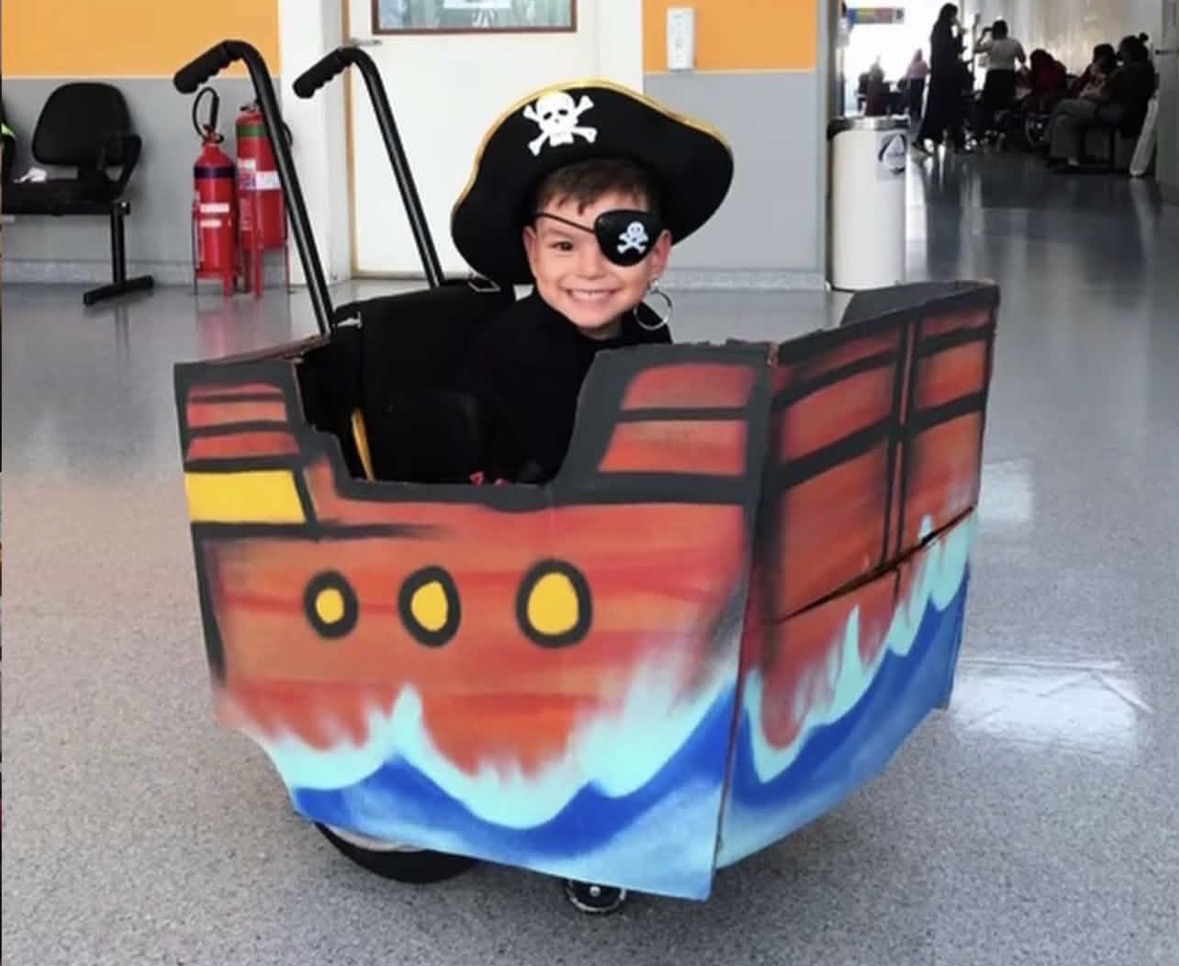 Otávio sorri com chapéu preto de pirata, tapa-olho e roupa preta, em cadeira de rodas cercada por uma estrutura de navio vermelho e marrom, com ondas azuis pintadas na base. Ao fundo, há cadeiras e pessoas em um corredor.
