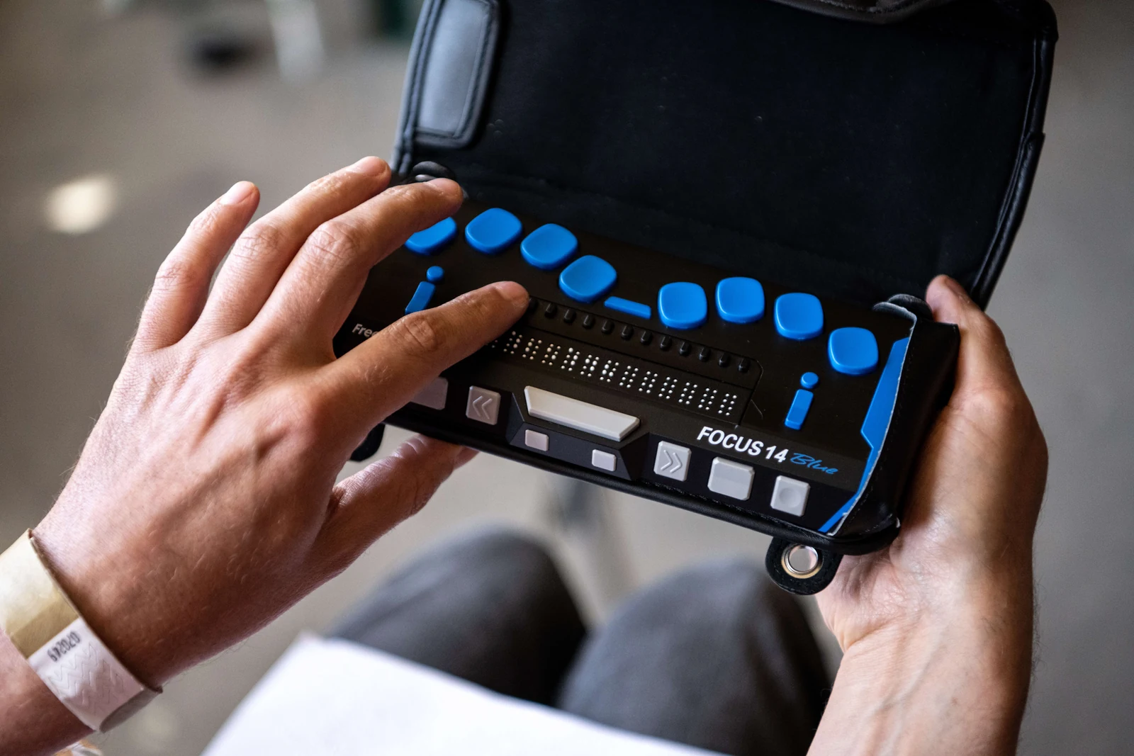 Mãos seguram e utilizam uma linha braille portátil preta, com teclas azuis e uma fileira de caracteres em relevo. Foto ilustrativa sobre tecnologia assistiva.