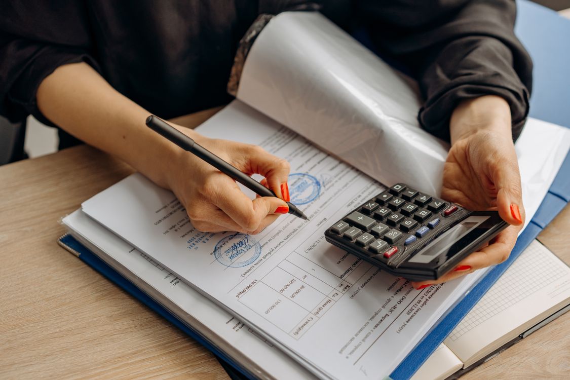 Mãos de uma pessoa analisam documentos e utilizam uma calculadora. Imagem ilustrativa sobre orçamento.