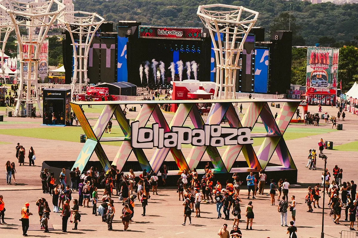 Visitantes circulam diante de um letreiro colorido com o nome Lollapalooza. Ao fundo, há um palco e outras estruturas do festival no Autódromo de Interlagos.