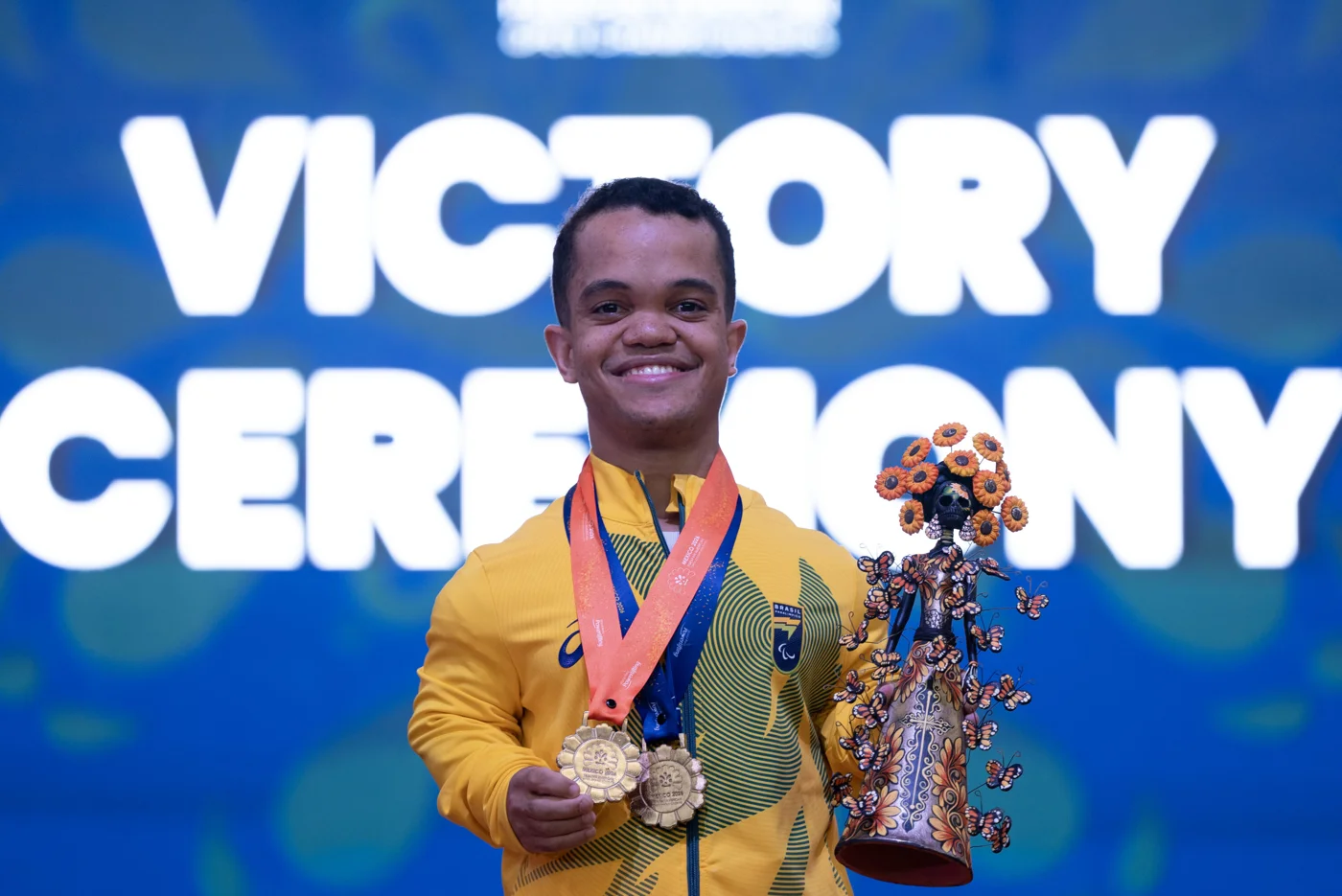 Marco Túlio Cruz sorri com uniforme amarelo do Brasil, medalhas no peito e uma escultura nas mãos durante a premiação no México.