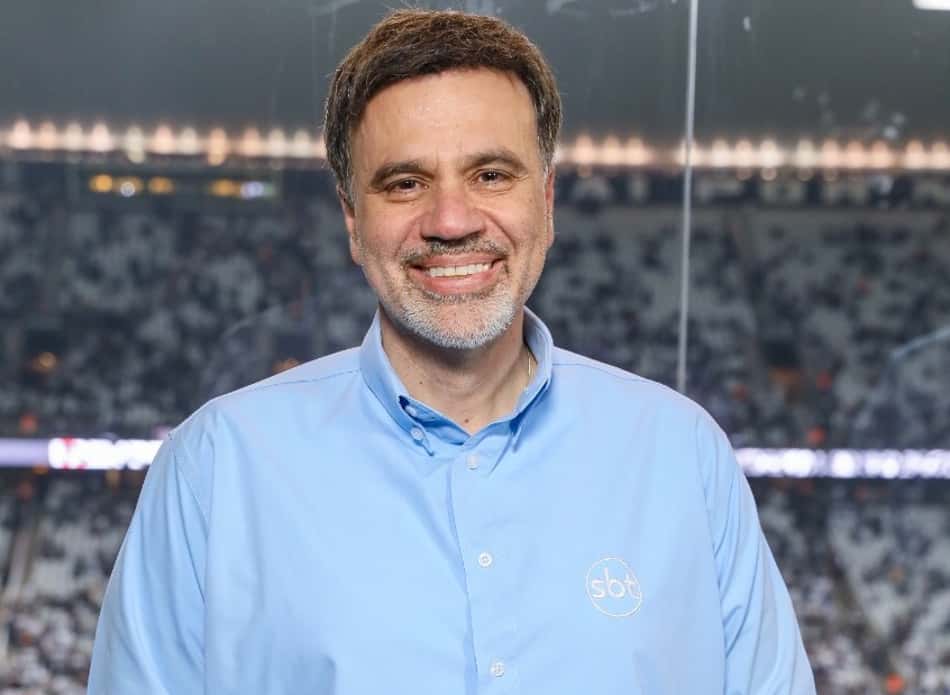 Mauro Beting sorri, usando camisa azul-clara com o logotipo do SBT. Ao fundo, aparecem as arquibancadas de um estádio de futebol.