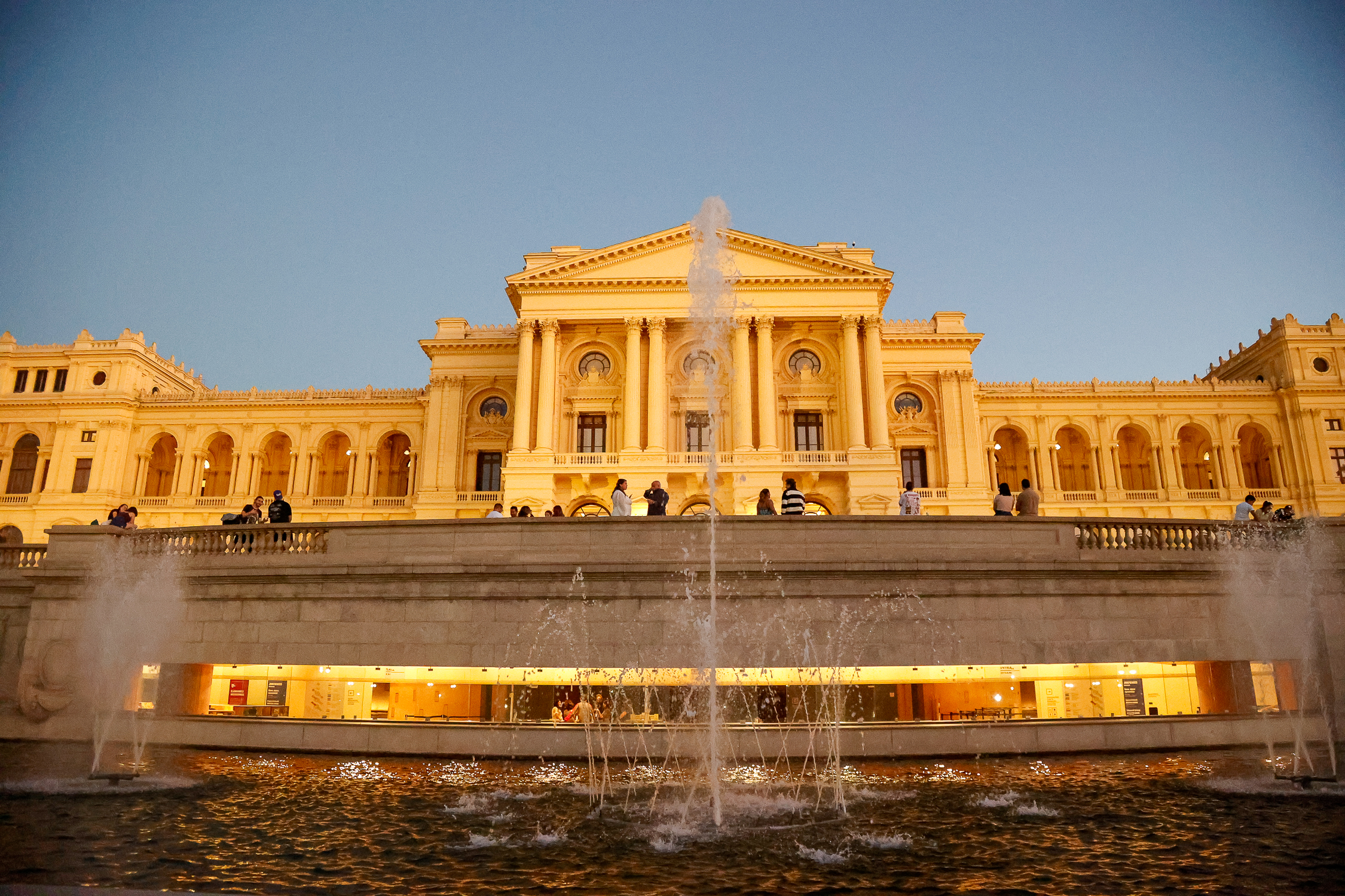 Fachada iluminada do Museu do Ipiranga, com fontes de água e visitantes ao entardecer.