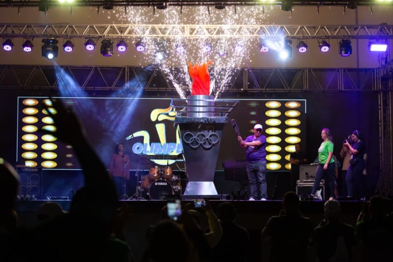 Pira acesa com faíscas em um palco da Olimpede, cercada por participantes, sob luzes azuis e roxas. Fotografia de arquivo.
