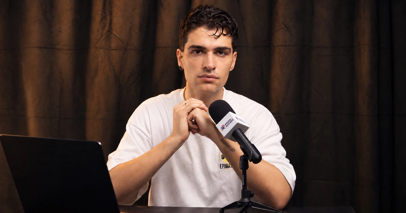 Victor Rodrigues, de camiseta branca, está sentado diante de um microfone do Modo de Acesso, com as mãos unidas. Um notebook aparece à esquerda e uma cortina escura ao fundo.