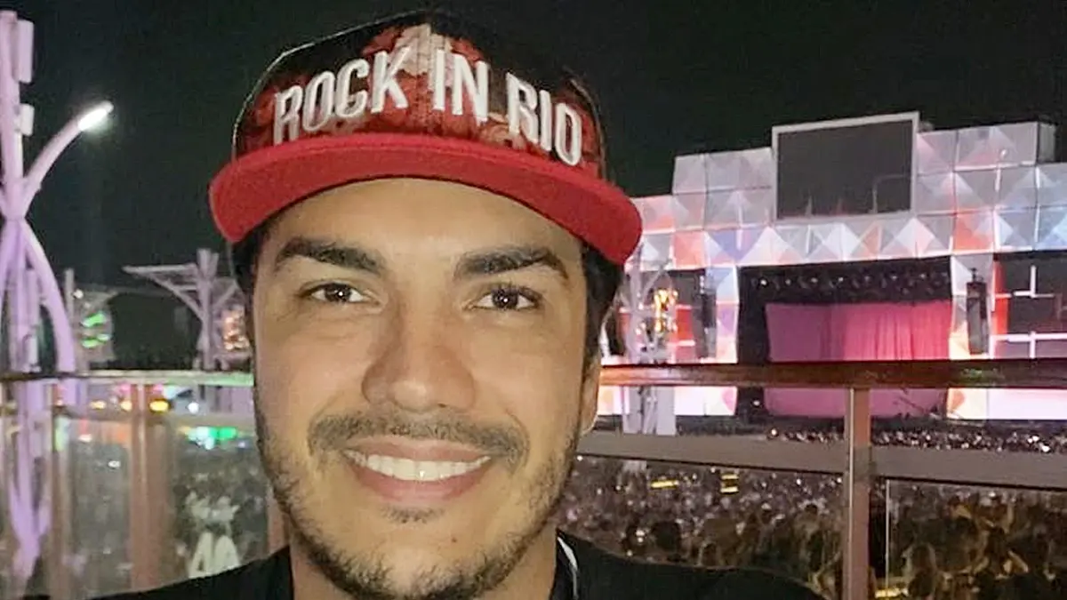 Thiago Amaral sorri para a câmera usando um boné vermelho com o nome Rock in Rio. Ao fundo, há um palco iluminado e o público do festival à noite.
